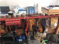 Full view of pegboard with various jars on top shelf, drill bits, clamps, protective masks, tape measures, tool bag, and other hand tools arranged on hooks and shelves.