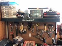 Wide view of black Mastercraft shelf with various tool storage boxes, tools hanging on pegboard below shelf including hammers, paintbrushes, wrenches, and other hand tools.
