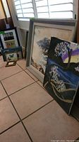 Several pieces of original artwork and paintings leaning against a wall and floor, showing various framed and canvas pieces, some signed, with visible styles including landscapes and floral themes.