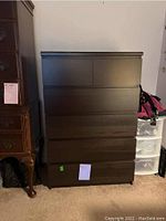 Full view of the dark wooden chest showing its front and side profile. The chest is placed next to other furniture and storage items with visible markings on the surface.