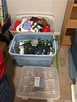 Three plastic bins shown containing Christmas decorations: fabric stockings, string lights with snowflakes, and boxed Hallmark Disney ornaments.