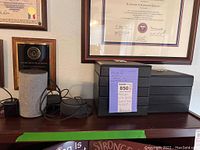 Photo showing Amazon Echo device, small round black device, and stacked black jewelry boxes with mirrored lids arranged on a dark wood surface under framed certificates.