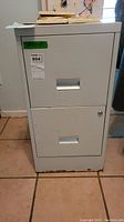 Front view of gray metal 2-drawer file cabinet showing recessed handles and lock on top drawer. Cabinet shows some rust at base.