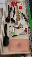 Photo of assorted kitchen utensils and food prep gadgets including silicone spatulas, metal whisk, measuring spoons, scissors, vegetable peeler, and plastic cutting board arranged in drawer.