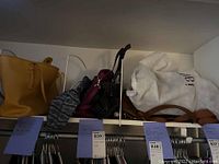 Photo showing a shelf with multiple handbags and purses including a yellow tote, patterned purse, black purse, maroon purse, and a white dust bag containing a handbag.