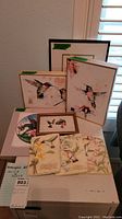 Overall view of hummingbird art collection including framed prints, embroidered panels, and decorative plate on white table.