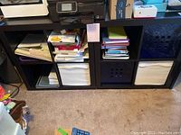Two IKEA cube storage units in dark wood finish, each with six compartments in 2x3 layout, containing black and white fabric bins and stacked papers/books. Located on carpeted floor with visible surface wear.