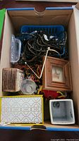 Boxes and costume jewelry strands in an open cardboard box, including decorative boxes and a picture frame