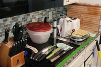 Wide angle photo showing knives in Sabatier Chef Star wooden block, Bobby Flay cutlery set, Cuisinart bagel toaster, Rapid egg cooker, wooden chopping block, plastic cutting board, and other kitchen tools on countertop.