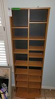 Front view of tall composite laminate media storage shelf with multiple compartments and wood-grain finish.