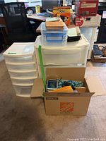 Overview of the lot showing plastic storage drawer units, cardboard box with office supplies and scissors, and small tote box.