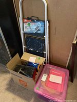 Folding stepstool with white metal frame and black steps showing paint splatters, two boxes containing assorted tools, hardware, and supplies.