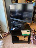 Wide view showing Westinghouse 32-inch TV on black TV stand with open shelves. Remote and other items on stand shelves and floor nearby.