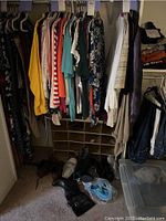 Closet view showing women's jackets, cardigans, and tops in colors such as yellow, teal, red, floral, and white hanging on hangers. Shoes and boots in multiple styles and colors are on the floor beneath the hanging clothes.