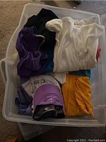 Top view of clear plastic bin containing folded activewear items including a white short sleeve top, black leggings, purple clothing, and a purple Nike visor.