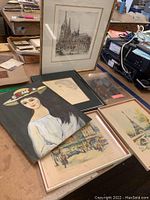 Photo showing six artworks including framed and unframed pieces on a table with various surroundings, depicting a black and white cathedral engraving, a portrait of a woman, and colorful cityscapes.