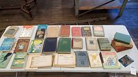 Top view of the table showing a large collection of vintage cookbooks and recipe books laid out in rows. Books vary in size, binding, and condition, with some colorful covers and some plain older ones.