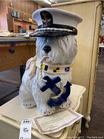 Front view of the ceramic dog figurine showing detailed navy cap with gold anchor, colorful collar with nautical flags, and large blue anchor held at front.