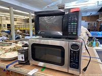 Top and front view showing black Goldstar MA795B microwave, stainless steel Sharp R-305KS microwave, and black Cuisinart DBM-8 coffee grinder on a table.