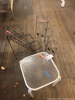 All five items in the lot including three black metal round plant stands, one black adjustable folding metal plant stand, and one glass top side table with white metal legs.