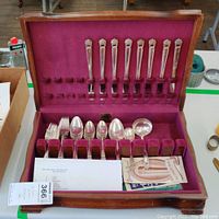 Open wooden box displaying multiple silver plated flatware pieces including forks, spoons and serving spoons arranged in velvet slots.
