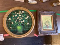 Pair of framed paintings on a dark wood surface, showing a round painting with daisies in a green jug and a rectangular floral oil painting in an ornate frame