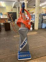 Front view of Oreck Classic XL vacuum upright with orange extension cord wrapped around handle, sitting on wooden floor in warehouse.