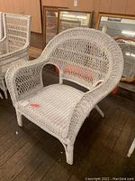 Photo of a large white wicker chair with rounded back and armrests, placed on a wooden floor with framed artwork visible in the background.