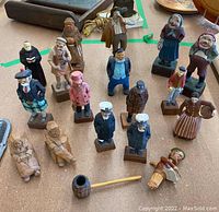 Overview of the 18 wooden carved figurines arranged on a table, showing diverse characters including sailors, men in kilts, elderly women, and a pipe among them.