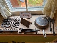 Full set of vintage cast iron kitchenware items displayed on a windowsill showing frying pan, molds, bacon presses, and serving spoons.