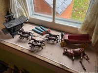 Black cast iron Crescent toy stove, wagon with six cast iron horses, cow bank, and blue pliers on fabric-covered window sill.