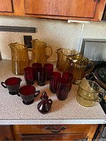View of the full set of amber glass pitchers, red drinking glasses, cream and sugar items, Avon cruet, and yellow glass bowls on countertop.