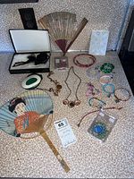 Lot overview showing various stone bead necklaces and bracelets, hand fans including a decorative fan with a painted woman, a white tissue pocket holder, and assorted costume jewelry