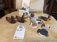 Full view of two wooden ducks, five resin ducks including one with Koreart sticker, ceramic egg ornament with duck painting, all displayed on a textile surface with tag "Ducks" and a white box behind.