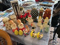 Wide view of 28 assorted figural candles on a table showcasing holiday and cherub themes, including white pillars, Santa Claus, snowmen, angels, and decorative pots.