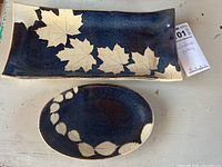 Two pottery trays shown side by side on a flat surface, one rectangular with leaf pattern, one oval with leaf motif.