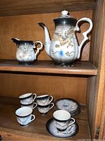 Full tea set arranged on wooden shelf, showing tea pot, creamer, six demitasse cups and saucers