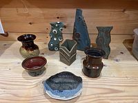 Seven pottery items on wooden surface: six vases in different shapes and colors, and one textured round plate with blue-black glaze.