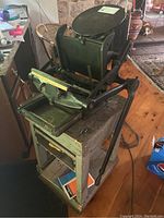 Manual black metal printing press on a painted wooden stand with peeling paint and slide-out tray. Visible mechanical lever arm and press plate, placed on wooden floor indoors.