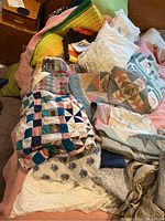 Photo showing various quilts, quilt tops, sheets and crochet throws laid out on a surface. Quilts have patchwork and geometric designs; several fabrics and patterns visible.