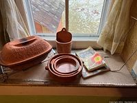 Romertopf clay pot with embossed lid darkened from use, metal handle stand, red ware baking dishes nested, two cookbooks on table by window.