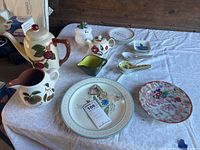 Full image of lot including Stangl pitcher, Franciscan teapot, green sauce pitcher, Asian spoons, and various plates on white fabric-covered surface
