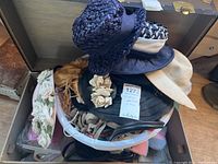 Box with various vintage hats stacked, showing different styles and materials, some with decorative flowers and bands, along with gloves, crochet slippers, and a glimpse of flapper dress fabric inside the trunk