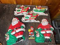 Six vintage-style plastic Santa Claus figures shown on a wooden surface. Five large Santas dressed in red with green mittens holding sacks of toys, along with three decorative sleighs - two red and one green.