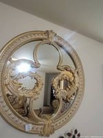 Front view of Anarina decorative wall mirror mounted on wall, showing gold ornate frame and intricate decorative scroll elements inside the mirror.