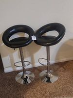 Two black bar stools side by side against a wall on carpeted floor, showing cushioned round seats with open backs and chrome pedestal bases with footrests.