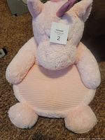 Front view of a pink plush unicorn chair for children, showing the textured seat and soft arms, feet, horn, and ears. The chair appears lightly used and in good condition.