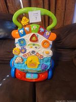 Front view of VTech Sit to Stand Learning Walker placed on a brown couch showing colorful interactive panel and green handle.