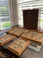 Photo showing four framed Navajo sand art pieces and the solid wood jewelry box. The art frames have native-inspired designs with sand in various colors, set in wooden frames. Jewelry box sits behind art pieces with double lattice screen doors and is medium wood tone.