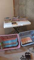 Multiple stacks of hardcover and softcover books on a folding table, plastic baskets on the floor contain numerous paperback books.